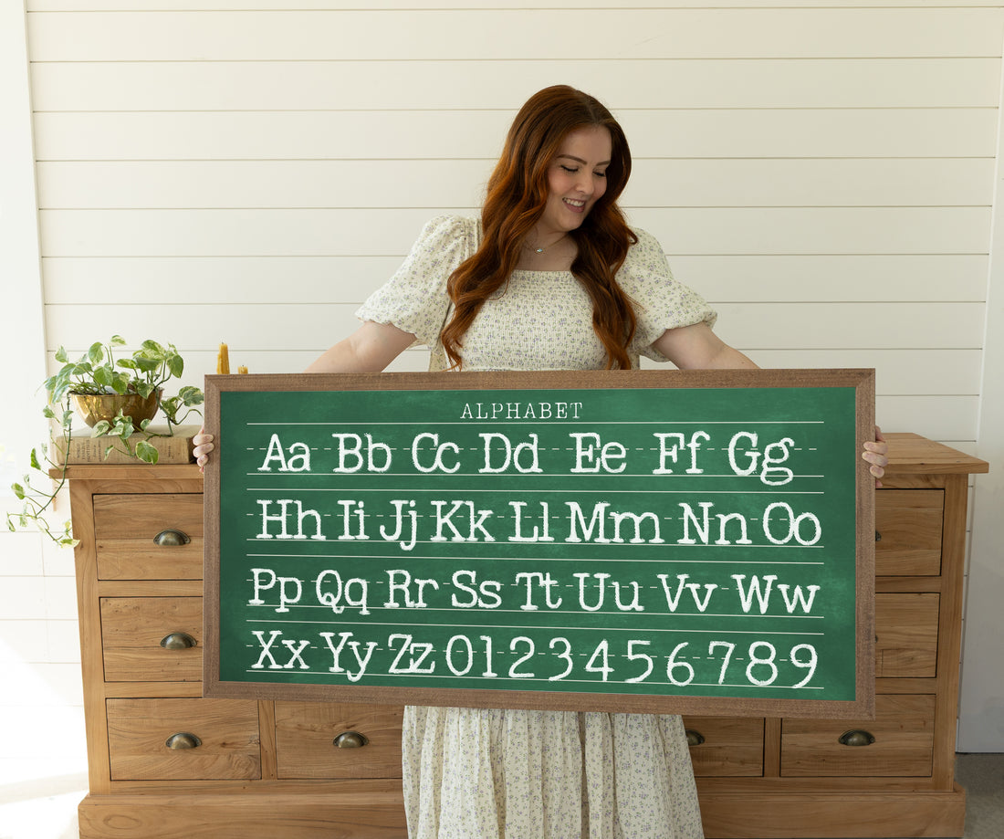 A woman is holding a large chalkboard with the alphabet written on it.